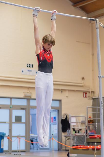Young male gymnast hangs from the high bar with intense focus, wearing red and black leo with white pants.