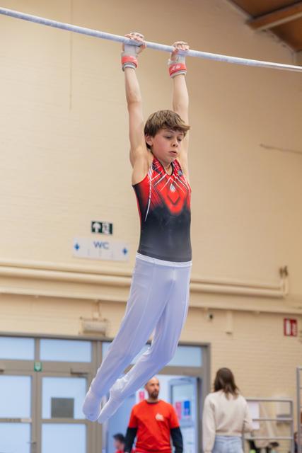 Young male gymnast hangs from the high bar with intense focus, wearing a red and black leotard with white pants.