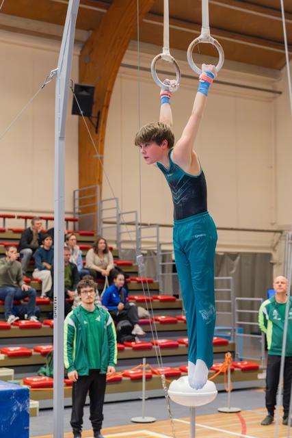 Young male gymnast hangs from still rings with focused expression, coach watching below in a gymnasium with spectators.