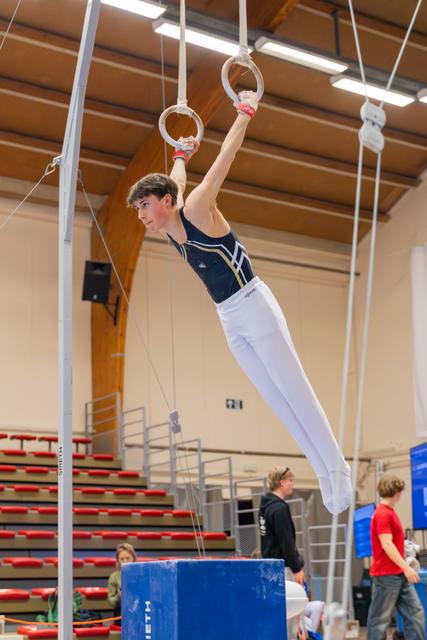 Young male gymnast hangs from still rings with focused expression, body angled, wearing navy leotard and white trousers.