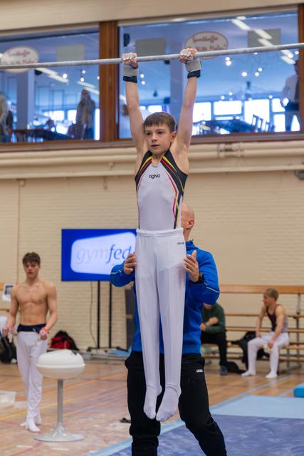 Young male gymnast grips the high bar with intense focus while a coach in blue steadies him from below in a gymnasium.