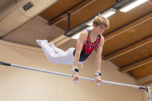 Young male gymnast grips the high bar with fierce focus, body horizontal, legs bent mid-routine in an indoor gymnasium.