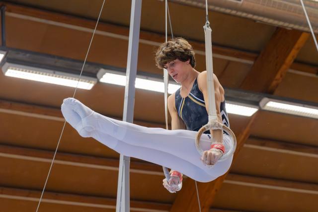 Young male gymnast holds a tuck position on still rings, focused and determined, inside a gymnasium.