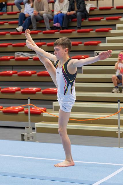 Young male gymnast balances on one leg with arms extended during a floor routine, focused and composed.