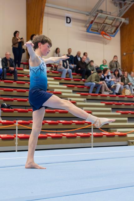 Young gymnast performs a precise leg raise on the floor mat, arms extended forward, focused and controlled, with spectators watching.