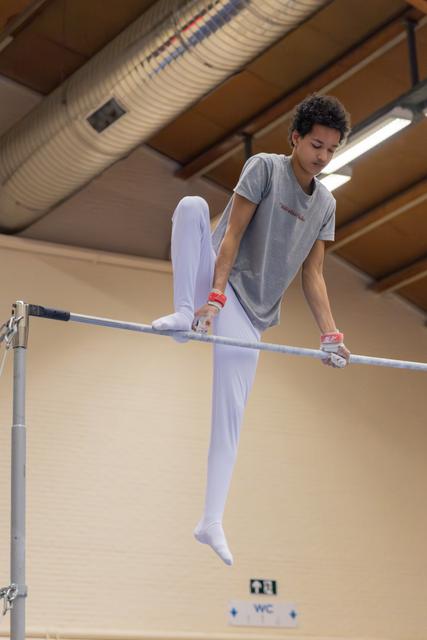 Young male gymnast grips parallel bars with focused concentration, legs mid-swing in an indoor gymnasium.