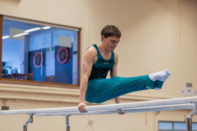 Young male gymnast performs a straddle L-sit on parallel bars, concentrating intensely in an indoor gymnasium.
