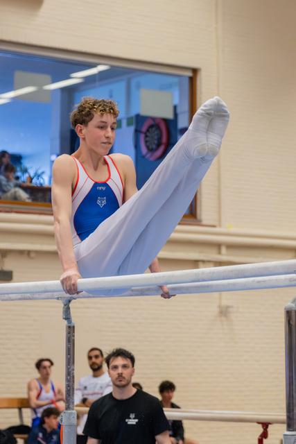 Focused teenage gymnast performs a straddle hold on parallel bars, legs raised high, wearing blue and white leotard.