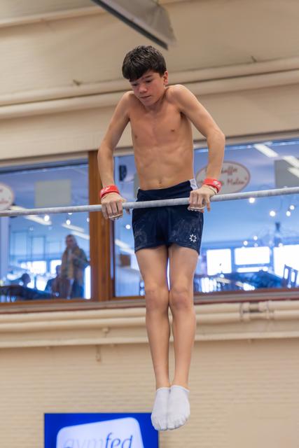 Shirtless teenage gymnast grips parallel bars with intense focus, body suspended mid-routine in an indoor gymnasium.