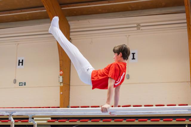Young boy in red shirt performs a leg raise on parallel bars, showing intense focus and strong body control in a gymnasium.