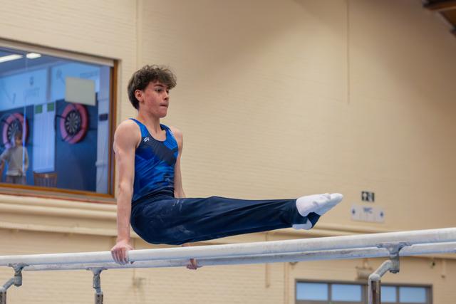 Focused teenage male gymnast holds a straddle L-sit on parallel bars, legs extended, expression intense and determined.