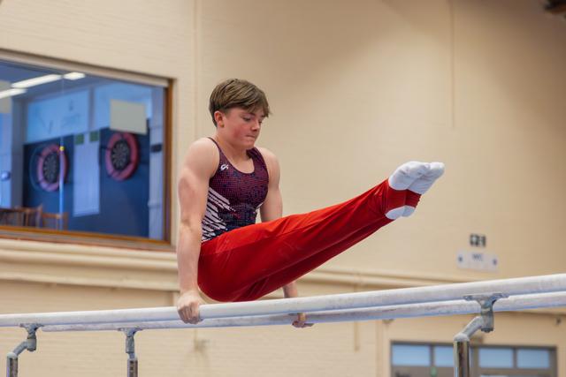 Young male gymnast performs a straddle hold on parallel bars, legs raised high, expression focused and intense indoors.