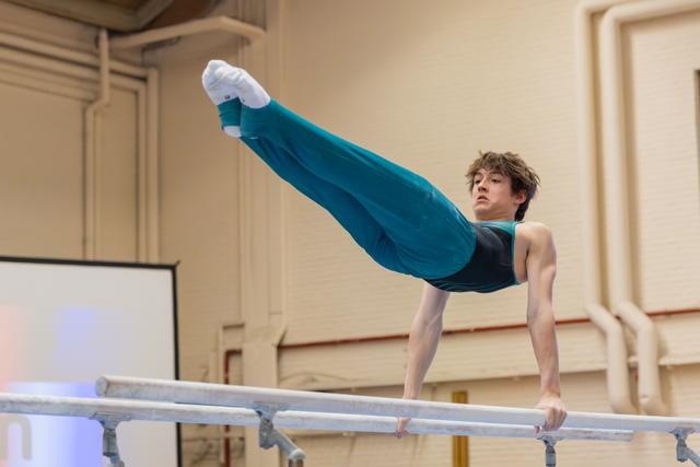 Young male gymnast performs a straddle hold on parallel bars, legs raised high, expression focused and determined.