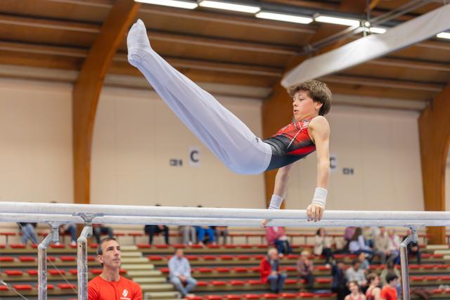 Young male gymnast performs a straddle hold on parallel bars, focused and controlled, as a coach watches below.