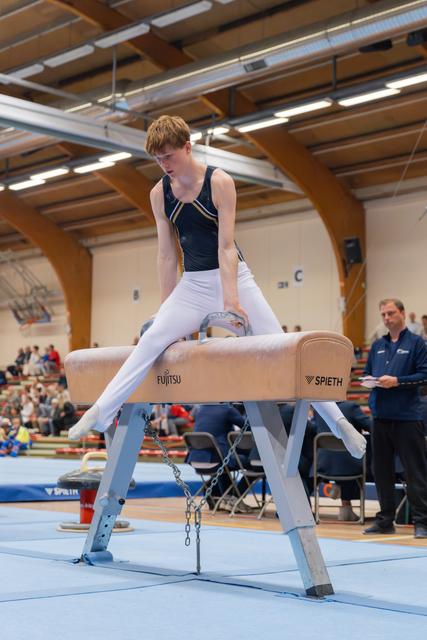 Focused teenage gymnast performs on a Spieth pommel horse during an indoor competition, wearing a dark blue leotard and white trousers.