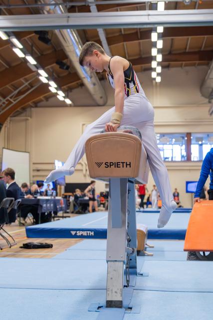 Young male gymnast leaning intently over a Spieth pommel horse, legs split mid-routine during an indoor gymnastics competition.