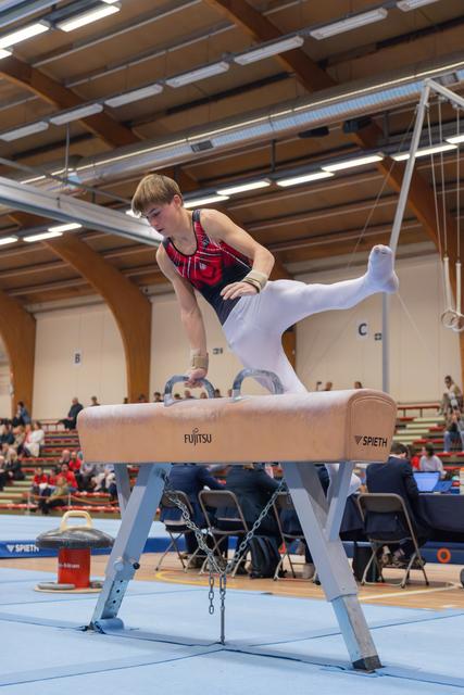 Young male gymnast performs on pommel horse, leaning forward with intense concentration, legs split mid-routine in indoor arena.