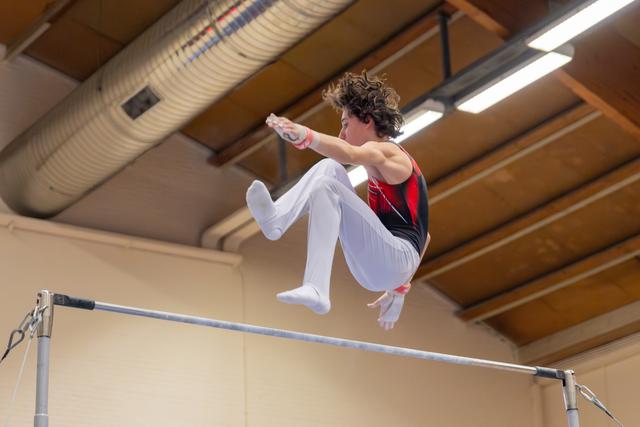 Young male gymnast releases the high bar mid-flight, tucking knees in a focused aerial dismount indoors.