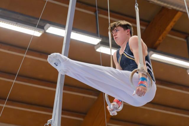 Young male gymnast performs on still rings, legs raised, focused expression, in an indoor gymnasium.