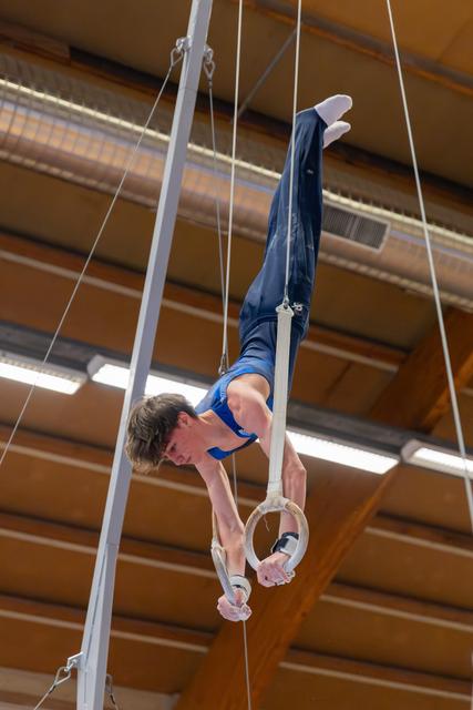 Young male gymnast performs an inverted pike hold on still rings, showing intense focus and strength in an indoor gymnasium.