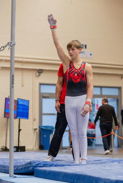 Young male gymnast raises one hand in salute before the high bar, composed and focused, coach standing behind him.