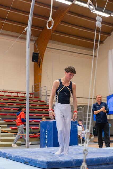 Young male gymnast stands composed on blue mat beneath still rings, coach applauding in background at indoor gymnastics meet.