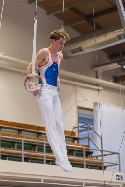 Young male gymnast hangs focused on still rings, wearing blue and white leotard in an indoor gymnastics hall.