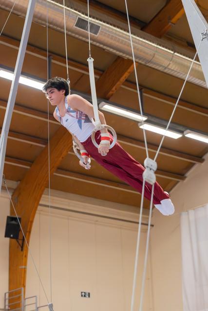 Young male gymnast performs on still rings, body horizontal, focused expression in a wooden-beamed gymnasium.