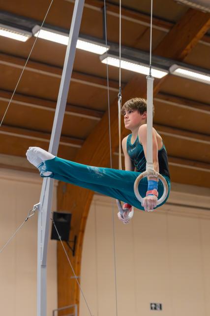 Young male gymnast performs on still rings, legs extended forward, focused expression in an indoor gymnastics hall.