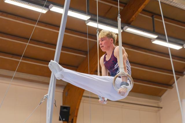 Young male gymnast performs on still rings, legs extended forward, intense concentration on his face, indoor gym setting.
