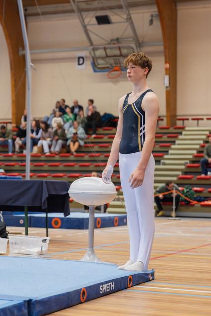 Teen male gymnast stands composed at chalk mushroom, focused expression, white leotard, gym competition hall with spectators.