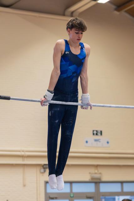 Teen male gymnast grips the high bar with intense focus, wearing a blue leotard and chalk-dusted grips.