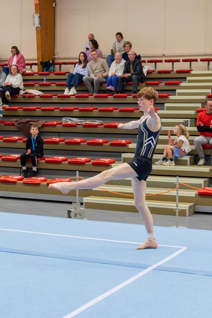 Teenage male gymnast performs a leg raise on the floor mat, focused and balanced, with spectators watching in the bleachers.