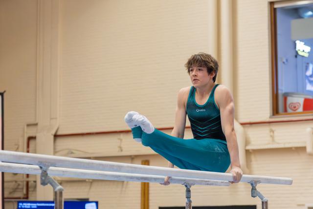 Focused teenage male gymnast holds an L-sit on parallel bars, legs extended forward, wearing teal leotard in indoor gym.