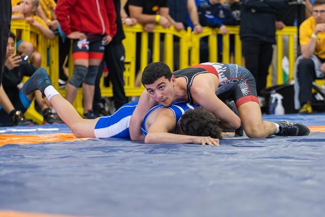 Young wrestler in black singlet pins opponent in blue, looking up confidently during an indoor match.