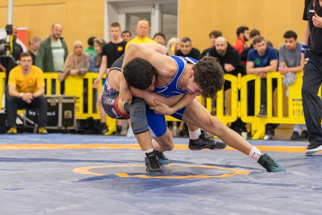 Wrestler in blue singlet drives opponent down with a powerful double-leg takedown on competition mat, crowd watching.