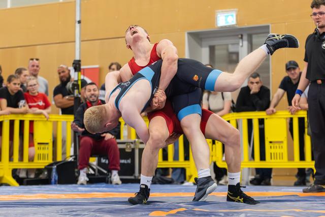 Wrestler in red grimaces as opponent in blue lifts him off the mat during an intense match before a packed crowd.