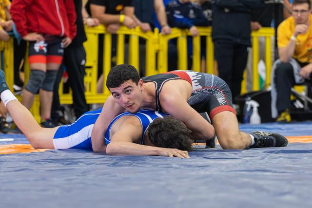 Young wrestler in black singlet pins opponent in blue, looking up confidently while controlling him on the mat.