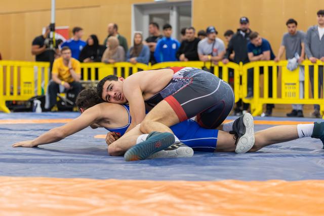 A wrestler in dark singlet pins his opponent to the mat, grimacing with effort, spectators watching behind yellow barriers.