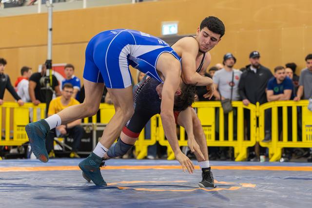 Wrestler in blue singlet drives opponent toward the mat in an aggressive takedown attempt on a blue competition floor.