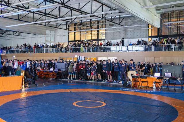 Young wrestlers stand at attention on the mat during an opening ceremony, watched by a packed crowd on the mezzanine above.