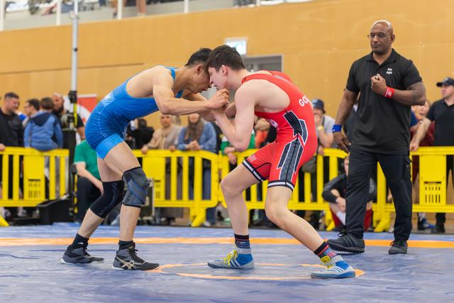 Two wrestlers in blue and red singlets lock grips on the mat, focused and tense, as a referee watches closely.