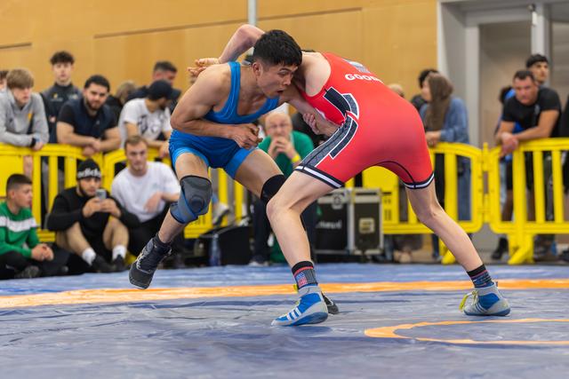 Two wrestlers clash on the mat, the blue-suited athlete lunging aggressively while the red-suited opponent braces firmly.