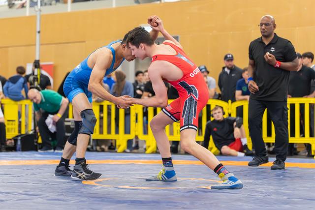 Two wrestlers grip hands in a tense standoff on the blue mat, referee watching closely from the sideline.