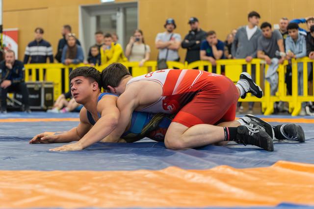 Young wrestler in blue singlet pinned down by opponent in red, struggling to escape on a blue competition mat.