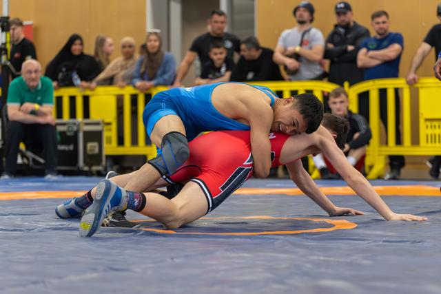 Wrestler in blue singlet aggressively controls opponent in red, driving them down onto the mat with intense focus.