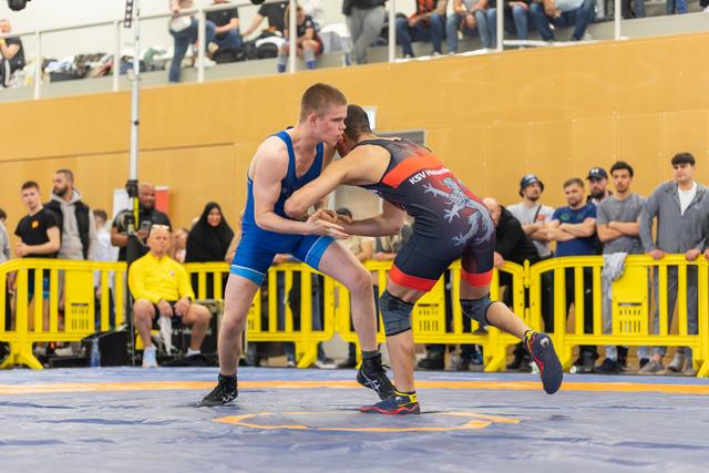 Two wrestlers locked in a grip battle on the mat, both in athletic stances, crowd watching intently behind yellow barriers.