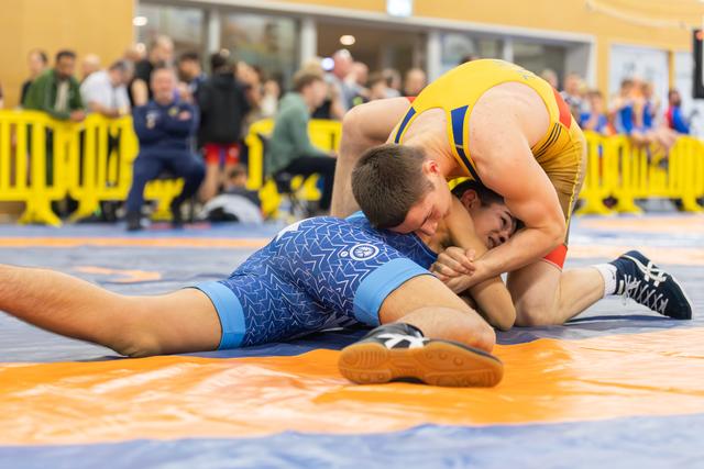 Wrestler in blue singlet pinned down by opponent in yellow, both straining intensely on orange and blue mat.
