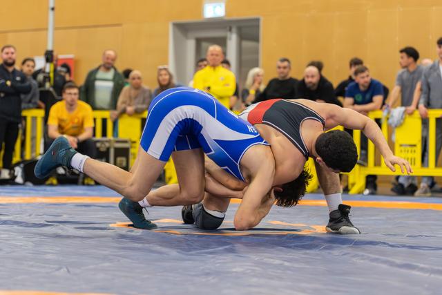 Two wrestlers grapple intensely on a blue mat, one in blue pinning his opponent down before a watching crowd.