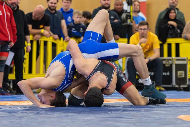 Two wrestlers grapple intensely on the mat, one in blue dominating with a takedown as spectators watch closely.
